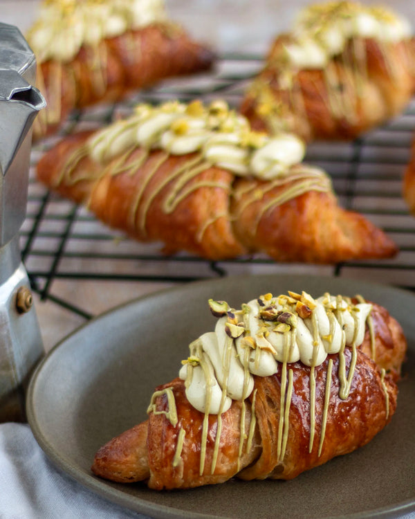 Honeywell Bakes Make Your Own Pistachio Croissant Baking Kit - Croissants with pistachio buttercream and pistachio drizzle on a plate, with more croissants on a cooling rack in the background.