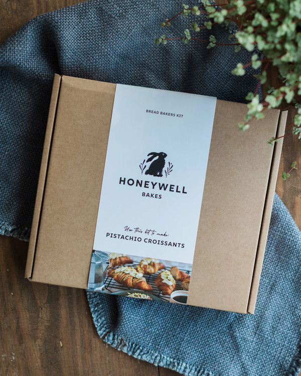 Honeywell Bakes Make Your Own Pistachio Croissant Baking Kit box on a wooden surface with a blue cloth and plant.