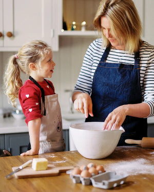 Honeywell Bakes Gingerbread House Baking Kit - A Mother and a young girl wearing aprons bake together in a bright kitchen. The woman in a striped shirt mixes ingredients in a large white bowl on a wooden table, with eggs, butter, and flour scattered around.