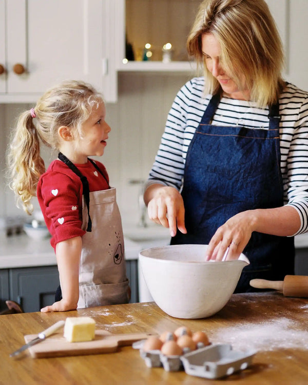 Honeywell Bakes Gingerbread House Baking Kit - A Mother and a young girl wearing aprons bake together in a bright kitchen. The woman in a striped shirt mixes ingredients in a large white bowl on a wooden table, with eggs, butter, and flour scattered around.