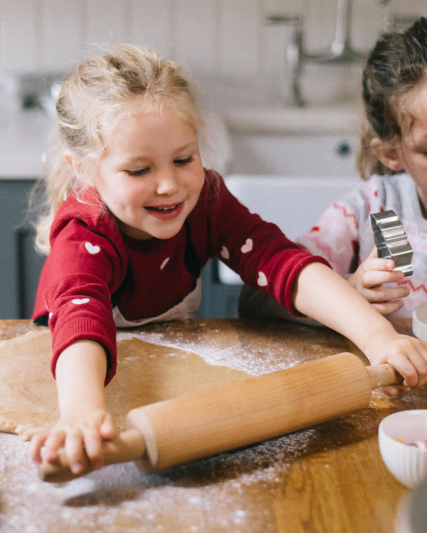 Honeywell Bakes Gingerbread House Baking Kit - A young girl with blonde hair wearing a red long-sleeve shirt with white hearts smiles while rolling out dough with a wooden rolling pin on a floured wooden table. Another child is partially visible on the right holding a metal cookie cutter.