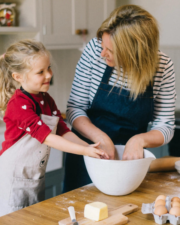 Honeywell Bakes Gingerbread House Baking Kit - A Mother and a young girl baking together in a kitchen, mixing ingredients in a large white bowl on a wooden table with butter, eggs, and flour.