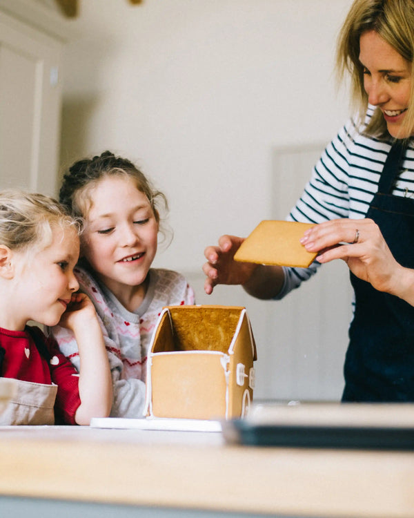 Honeywell Bakes Gingerbread House Baking Kit - A Mother and two children are gathered around a gingerbread house on a table in a home setting. The woman, wearing a navy apron over a black and white striped shirt, holds a piece of gingerbread to attach to the house.
