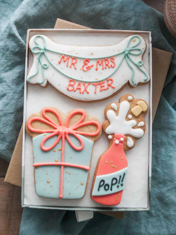 Honeywell Bakes - Medium Happy Birthday Biscuit Gift Set - Decorative cookies shaped like a gift box and champagne bottle with 'MR & MRS BAXTER' on a blue fabric background.