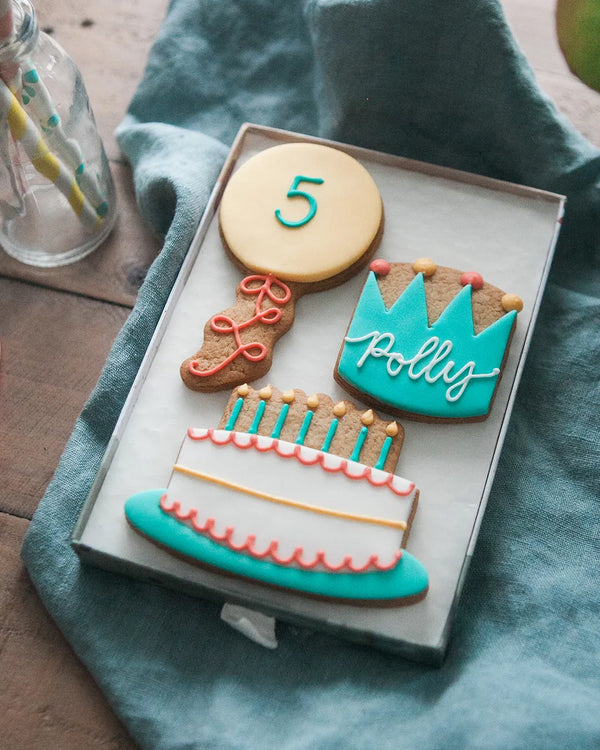 Honeywell Bakes Medium Birthday Party Biscuit Gift- Decorative cookies shaped like a balloon with a number 5 piped on, cake, and crown with personalised name on a blue cloth.