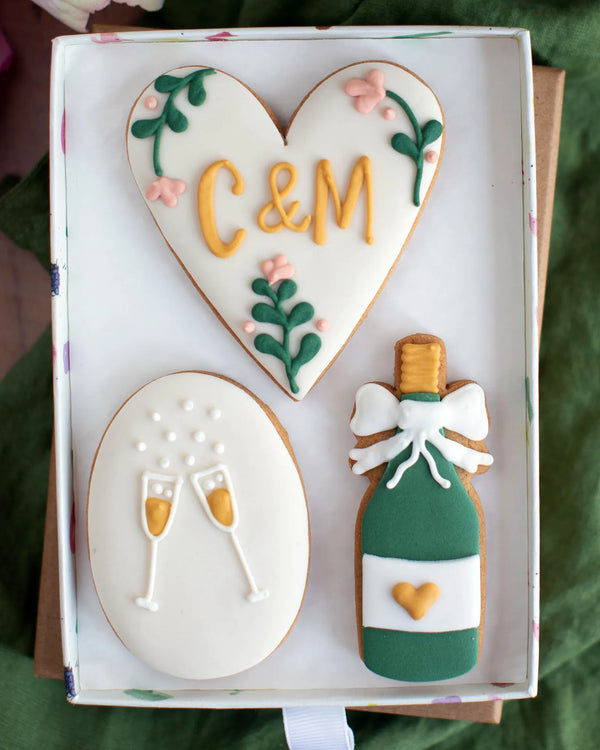 Honeywell Bakes Medium Couple Celebration Biscuit Gift Set - Decorative hand iced cookies shaped like a heart with personalised initials', a bottle of champagne, and champagne glasses in a gift box.