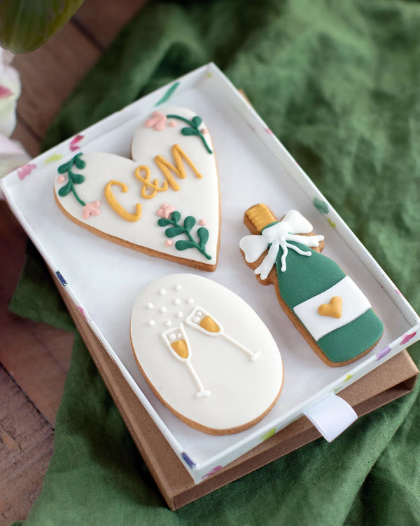 Honeywell Bakes Medium Couple Celebration Biscuit Gift Set - Decorative hand iced cookies shaped like a heart with personalised initials', a bottle of champagne, and champagne glasses in a gift box.
