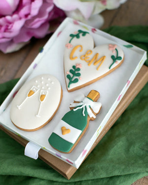 Honeywell Bakes Medium Couple Celebration Biscuit Gift Set - Decorative hand iced cookies shaped like a heart with personalised initials', a bottle of champagne, and champagne glasses in a gift box.