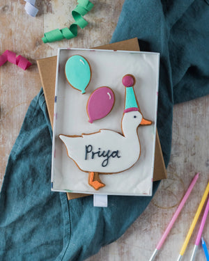 Honeywell Bakes Medium Party Duck Biscuit Gift - Decorative cookie with 'Priya' on a wooden surface with colourful ribbons and pens.