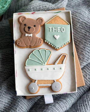 Honeywell Bakes Medium Welcome Baby Biscuit Gift - Three decorated cookies shaped like a teddy bear, a baby carriage, and a personalised sign in a gift box on a textured surface.