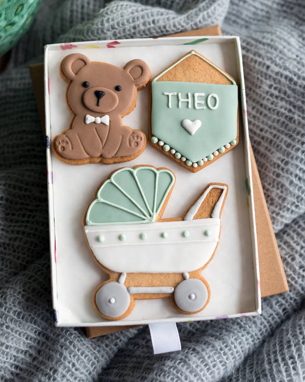 Honeywell Bakes Medium Welcome Baby Biscuit Gift - Three decorated cookies shaped like a teddy bear, a baby carriage, and a personalised sign in a gift box on a textured surface.