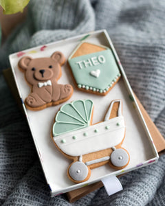 Honeywell Bakes Medium Welcome Baby Biscuit Gift - Three decorated cookies shaped like a teddy bear, a baby carriage, and a personalised sign in a gift box on a textured surface.