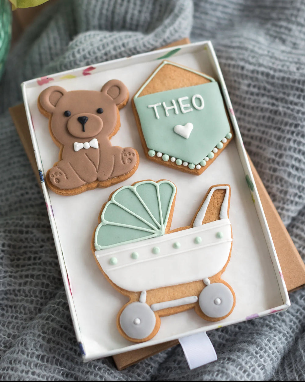 Honeywell Bakes Medium Welcome Baby Biscuit Gift - Three decorated cookies shaped like a teddy bear, a baby carriage, and a personalised sign in a gift box on a textured surface.