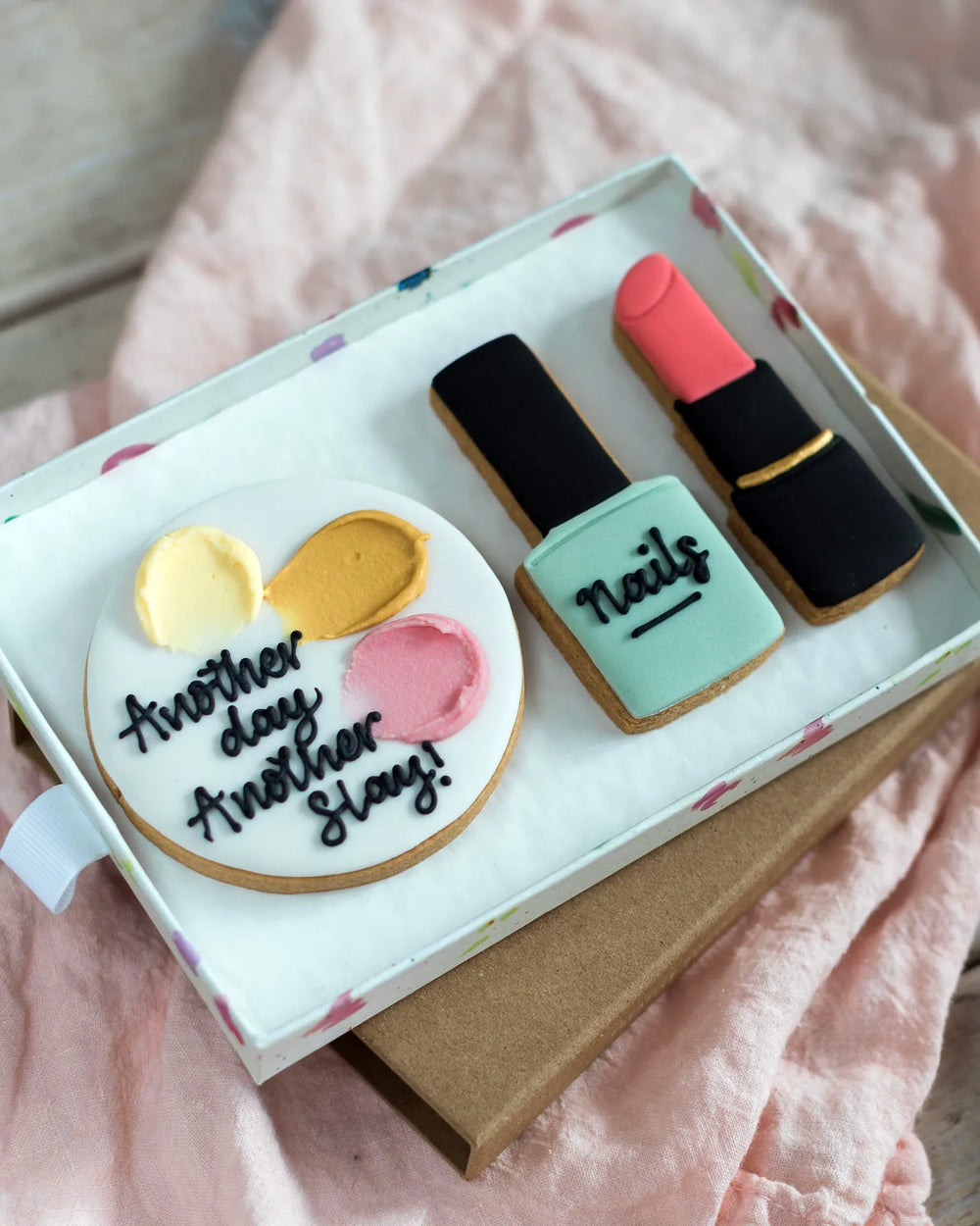 Honeywell Bakes Medium Beauty Biscuit Gift Set - Cookie set shaped like a birthday cake, nail polish bottle, and lipstick in a box on a  pink surface.