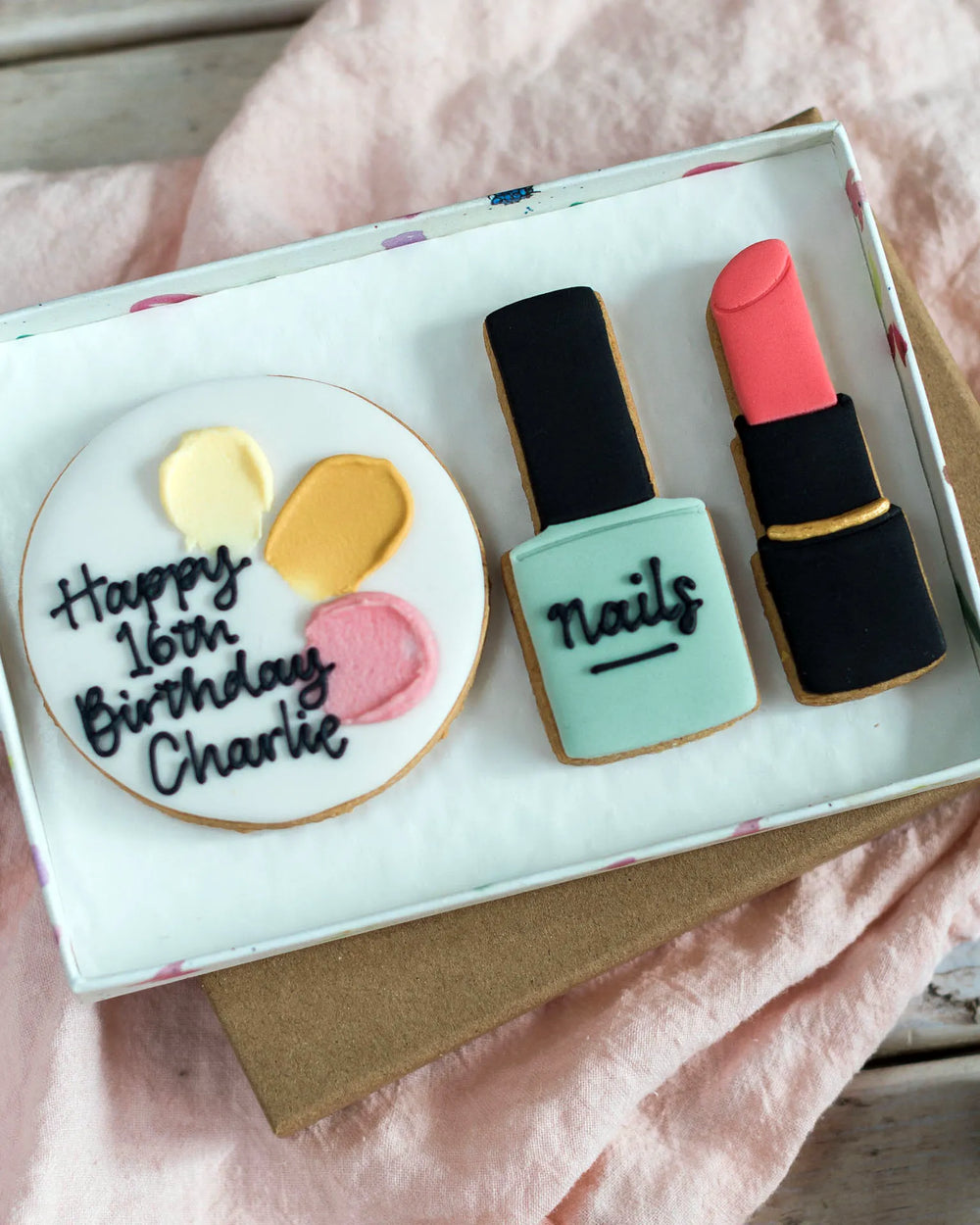 Honeywell Bakes Medium Beauty Biscuit Gift Set - Cookie set shaped like a birthday cake, nail polish bottle, and lipstick in a box on a  pink surface.