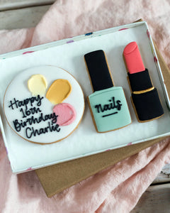 Honeywell Bakes Medium Beauty Biscuit Gift Set - Cookie set shaped like a birthday cake, nail polish bottle, and lipstick in a box on a  pink surface.