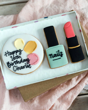 Honeywell Bakes Medium Beauty Biscuit Gift Set - Cookie set shaped like a birthday cake, nail polish bottle, and lipstick in a box on a  pink surface.