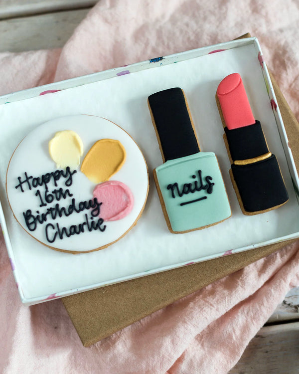 Honeywell Bakes Medium Beauty Biscuit Gift Set - Cookie set shaped like a birthday cake, nail polish bottle, and lipstick in a box on a  pink surface.