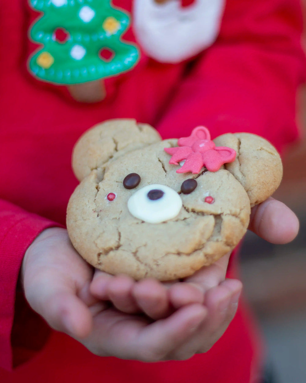 Honeywell Bakes - Four decorated teddy bear-shaped cookies in a cardboard tray on a wooden table, with additional cookies on plates and Christmas tree branches and lights in the background.