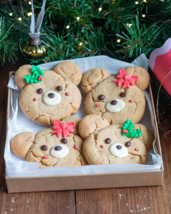 Honeywell Bakes - Four teddy bear-shaped cookies decorated with white frosting faces, chocolate chip eyes and noses, and colourful icing holiday accents, presented in a brown cardboard box lined with white tissue paper on a wooden table with a Christmas tree in the background.