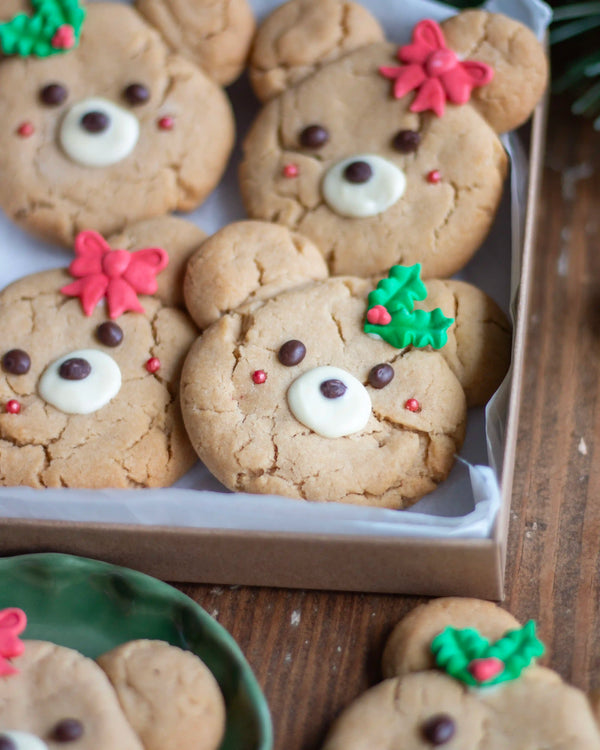 Honeywell Bakes - Decorated holiday teddy bear cookies arranged in a tray on a wooden table, with icing faces, chocolate eyes, and red and green icing accents.