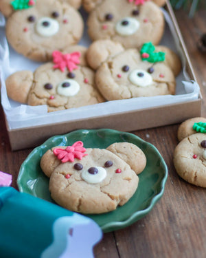 Honeywell Bakes - Decorated teddy bear-shaped cookies with white and brown frosting eyes, pink bow accents, and green leaf details, shown in a box and on a green plate on a wooden table.