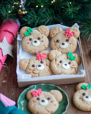 Honeywell Bakes - Four decorated teddy bear-shaped cookies in a cardboard tray on a wooden table, with additional cookies on plates and Christmas tree branches and lights in the background.