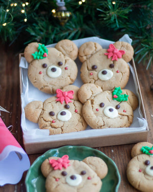 Honeywell Bakes - Four decorated teddy bear-shaped cookies in a cardboard tray lined with white parchment paper, set on a wooden table with Christmas greenery and lights in the background.