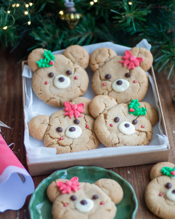 Honeywell Bakes - Four decorated teddy bear-shaped cookies in a cardboard tray lined with white parchment paper, set on a wooden table with Christmas greenery and lights in the background.