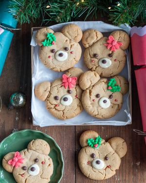 Honeywell Bakes - Four decorated bear cookies arranged on a tray with white parchment paper, surrounded by holiday greenery, wrapped gifts, and a green plate on a dark wooden table.