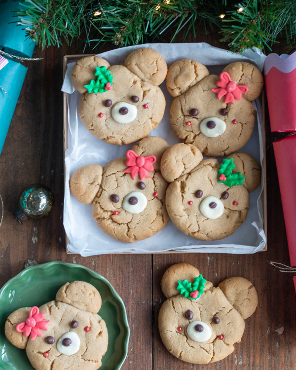 Honeywell Bakes - Four decorated bear cookies arranged on a tray with white parchment paper, surrounded by holiday greenery, wrapped gifts, and a green plate on a dark wooden table.