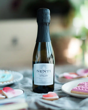 Honeywell Bakes Mini Bottle of Prosecco - A bottle of Senti Extra Dry sparkling wine with a black foil top and white label, set on a table with blurred heart-shaped cookies and a soft-focus background.