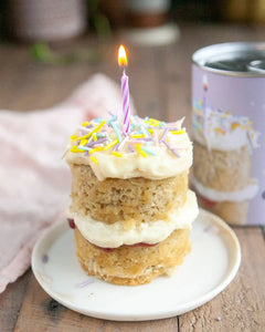 Honeywell Bakes Mini Celebration Cake Kit - Small birthday cake with a lit candle on a white plate, with a blurred background.