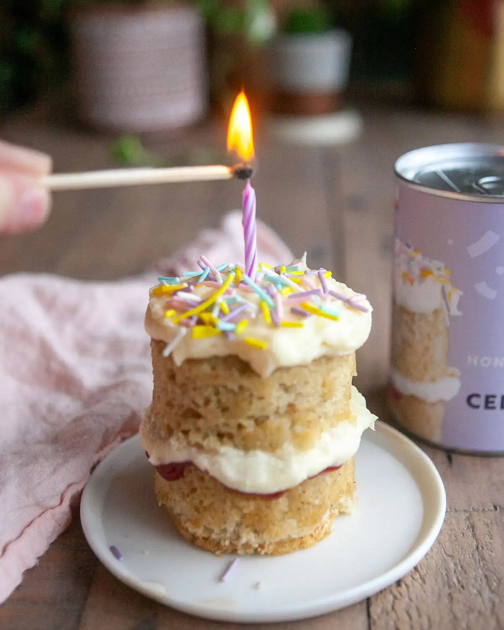 Honeywell Bakes Mini Celebration Cake Kit - Small birthday cake with a lit candle on a white plate, with a blurred background.