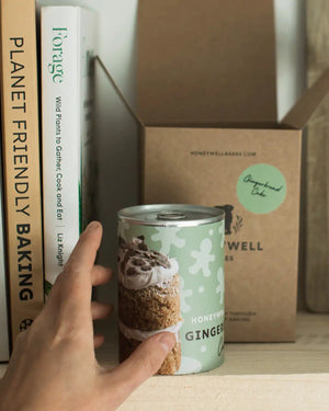 Honeywell Bakes Mini Gingerbread Cake Kit - Hand holding a can of ‘Honeywell Bakes Mini Gingerbread Cake' against a background with books and a box.