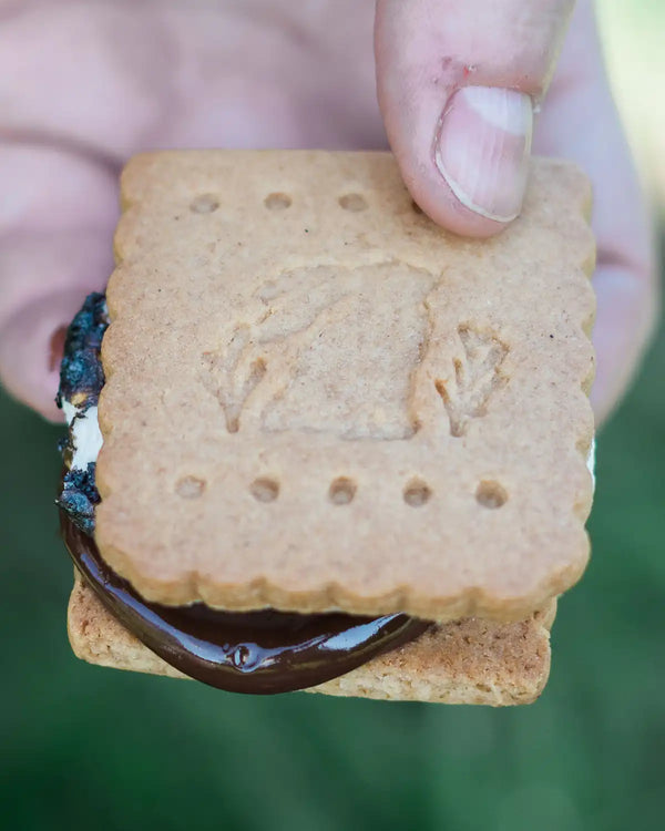 Honeywell Bakes Mini S’mores Kit - Hand holding a s'mores cookie with chocolate filling against a blurred green background