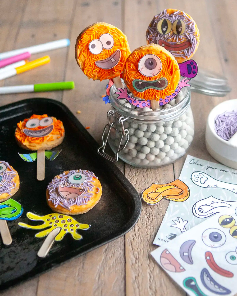 Honeywell Bakes Monster Biscuit Pop Baking Kit - Colourful monster-themed cookies on sticks with activity card and decorations on a wooden table.