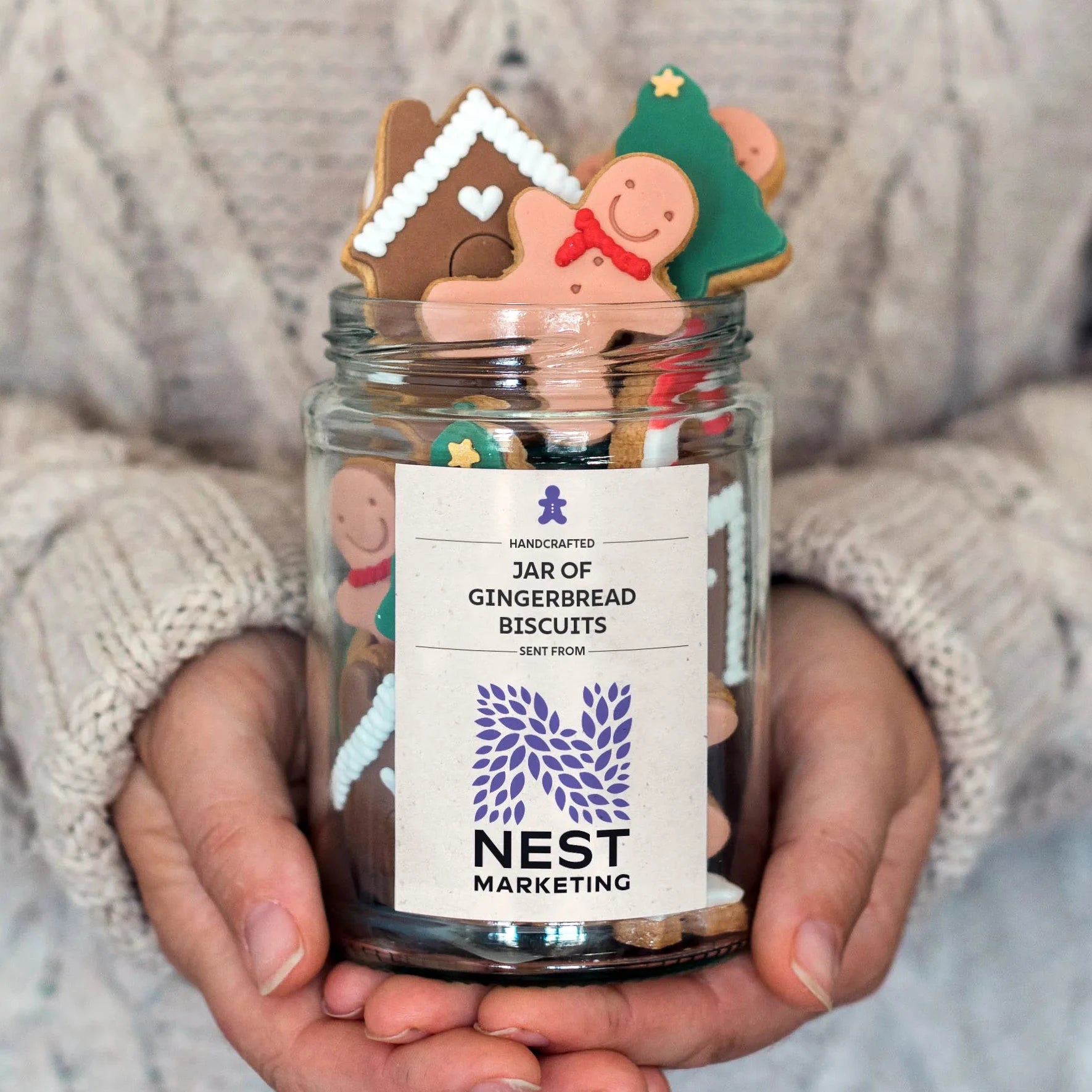 Honeywell Bakes Personalised branded gingerbread jar; A person in a cream cable-knit sweater holds a clear glass jar filled with gingerbread cookies. The white label on the jar reads 'HANDCRAFTED JAR OF GINGERBREAD BISCUITS' and shows the brand 'NEST MARKETING'.