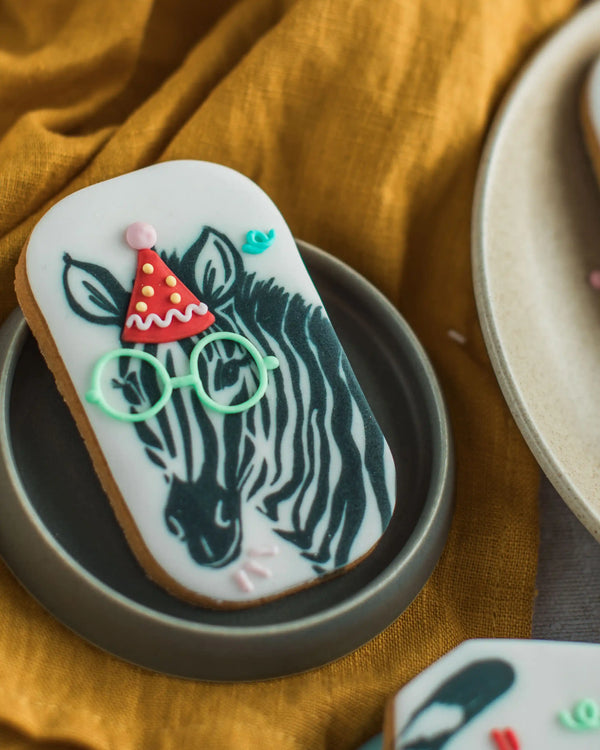 Honeywell Bakes Party Animal Biscuit Gift - Decorative cookie with a zebra wearing glasses and a party hat on a yellow fabric background.