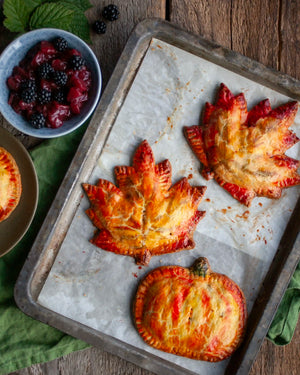 Honeywell Bakes - Three baked maple leaf and pumpkin hand pies on a parchment-lined baking tray, with a bowl of fresh blackberries and part of another pie visible on a rustic wooden table.