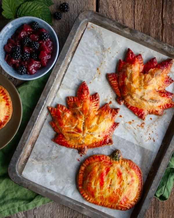 Honeywell Bakes - Three baked maple leaf and pumpkin hand pies on a parchment-lined baking tray, with a bowl of fresh blackberries and part of another pie visible on a rustic wooden table.