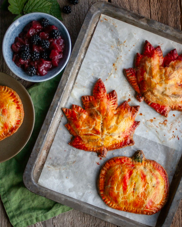 Honeywell Bakes - Three baked maple leaf and pumpkin hand pies with red berry filling sit on a parchment-lined baking tray, alongside a bowl of fresh blackberries and a green cloth on a rustic wooden table.