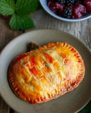 Honeywell Bakes A baked hand pie with a golden-brown crust sits on a gray plate on a rustic wooden table, with a bowl of mixed berries and green leaves in the background.