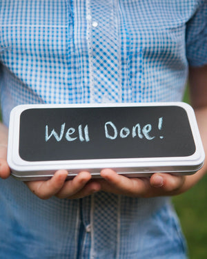 Honeywell Bakes Personalised Biscuit Pencils in Pencil Tin - Person holding a small chalkboard with 'Well Done!' written on it, wearing a blue checkered shirt.