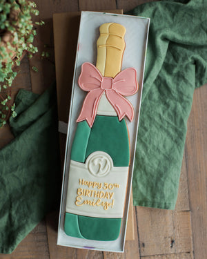 Honeywell Bakes Personalised Champagne Biscuit Bottle - Green - Cookie shaped like a champagne bottle with a pink bow and 'Happy 50th Birthday Emily!' text, presented in a box on a wooden surface.