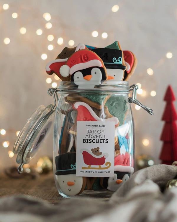 Honeywell Bakes Personalised Christmas Advent Calendar Biscuit Jar - A clear glass jar filled with assorted Christmas-themed biscuits, topped with penguin and Santa cookie cutouts, with a hanging tag reading 'Jar of Advent Biscuits' and warm blurred fairy lights in the background.