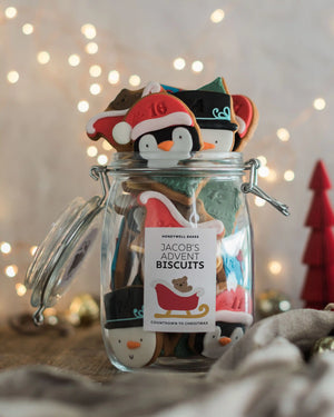 Honeywell Bakes Personalised Christmas Advent Calendar Biscuit Jar - A clear glass jar filled with Christmas-themed cookies, topped with penguin and Santa cookies, and labeled 'JACOB'S ADVENT BISCUITS'. The jar is set on a rustic surface with warm bokeh string lights in the background.