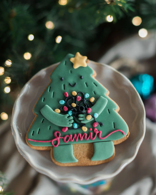 Personalised Christmas Tree Biscuit - A Christmas tree-shaped sugar cookie decorated with teal icing, a gold star, colourful candy-cane ornaments, and the name ‘Samir' in pink script, served on a white plate with a blurred festive background.