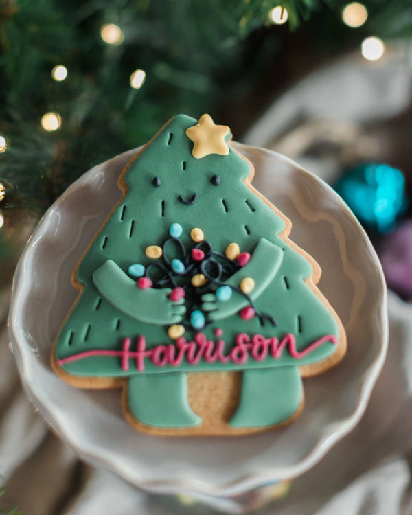Personalised Christmas Tree Biscuit - A Christmas tree-shaped sugar cookie decorated with green icing, colourful lights, a gold star, and the name 'Harrison' in pink script, served on a white plate with a blurred holiday background.