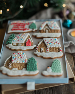 Honeywell Bakes Personalised Christmas Village Biscuit Gift - Six decorated gingerbread house cookies arranged on a light blue tray, with festive blurred holiday lights and ornaments in the background.
